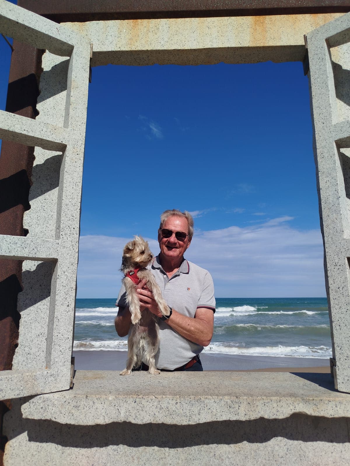 Martin Feijen holding a small dog by a stone structure with a beach backdrop